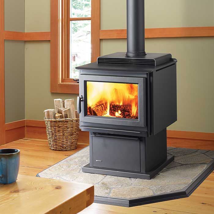 Wood burning standalone stove in black sitting on a stone hearth basket of wood to the left