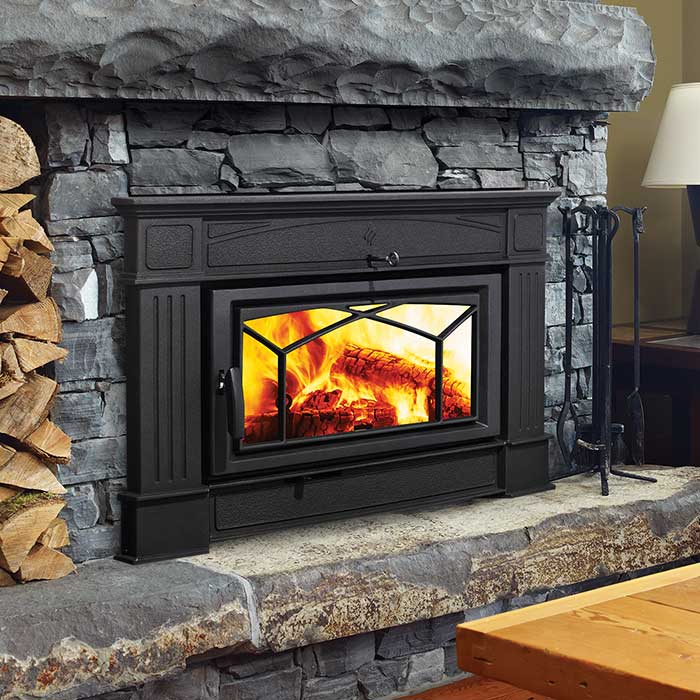 Wood burning insert with black facing and chinked stone mantle and stone surround with wood stacked to the left and tools on the right